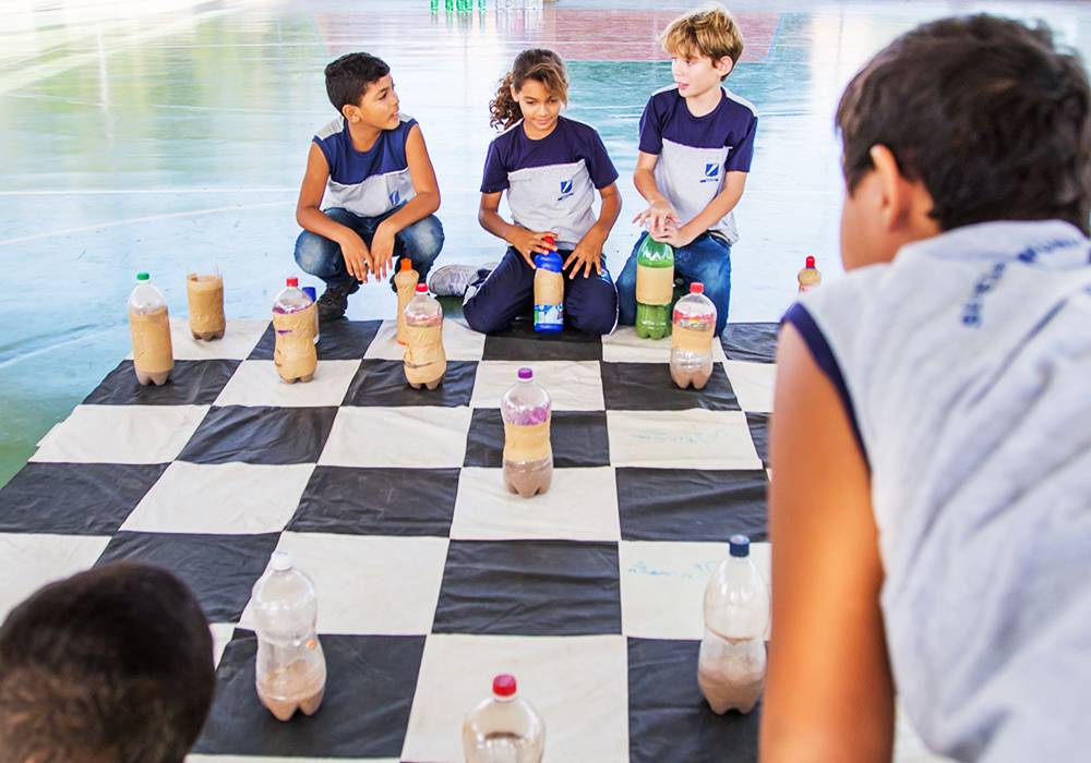 Três crianças analisam tabuleiro gigante de xadrez com peças feita de garrafa PET.