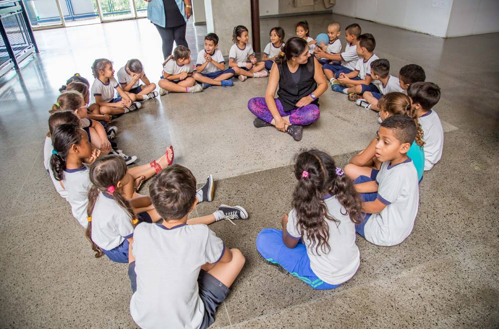 Meninas e meninos sentados no chão em roda com professora sentada ao centro.