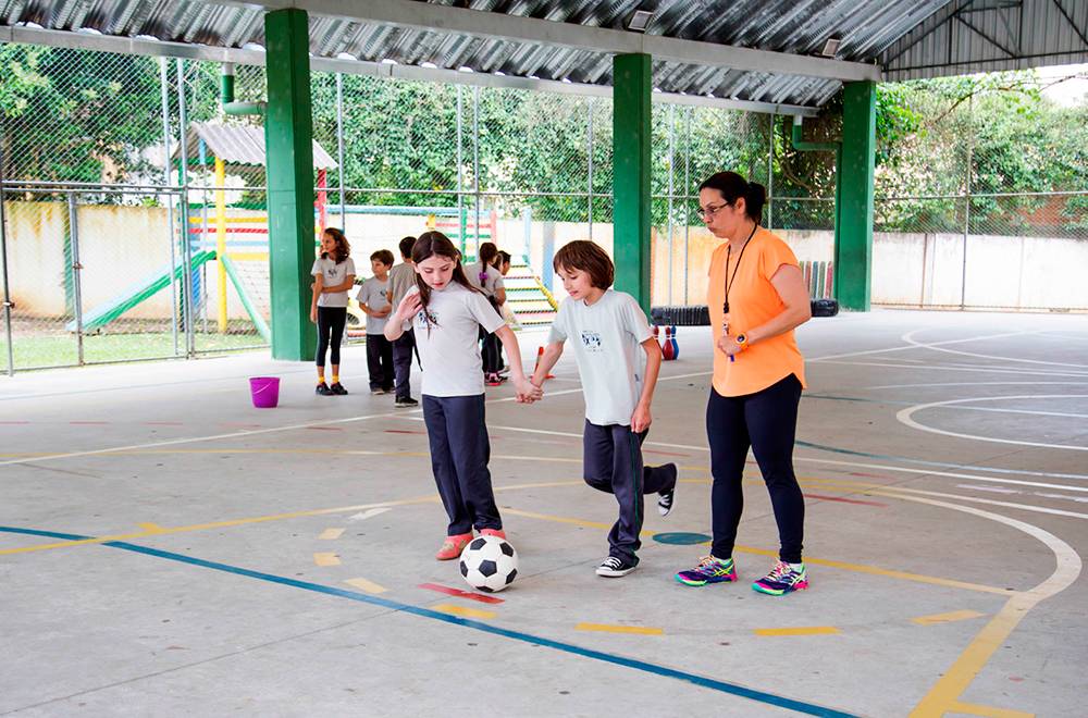 Menino prepare-se para chutar bola de futebol, ao lado uma colega segura sua mão enquanto professora a observa.