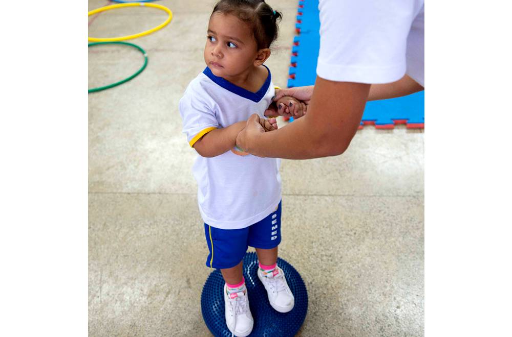 Menina se equilibra em bola pequena com a ajuda da professora.