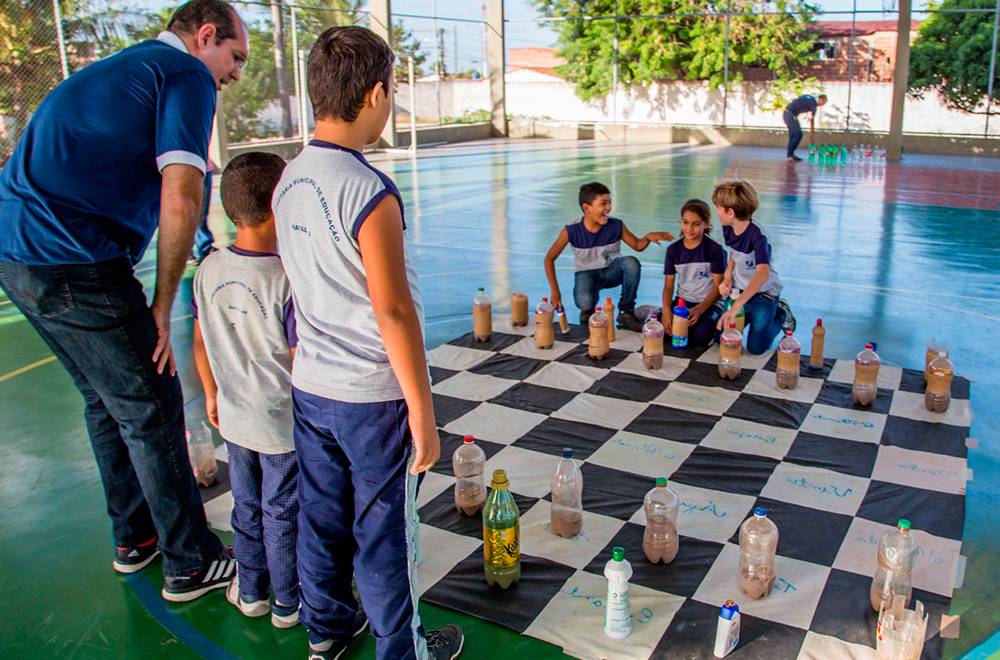 2 grupos de estudantes analisam jogo de xadrez em lados opostos do tabuleiro gigante.
