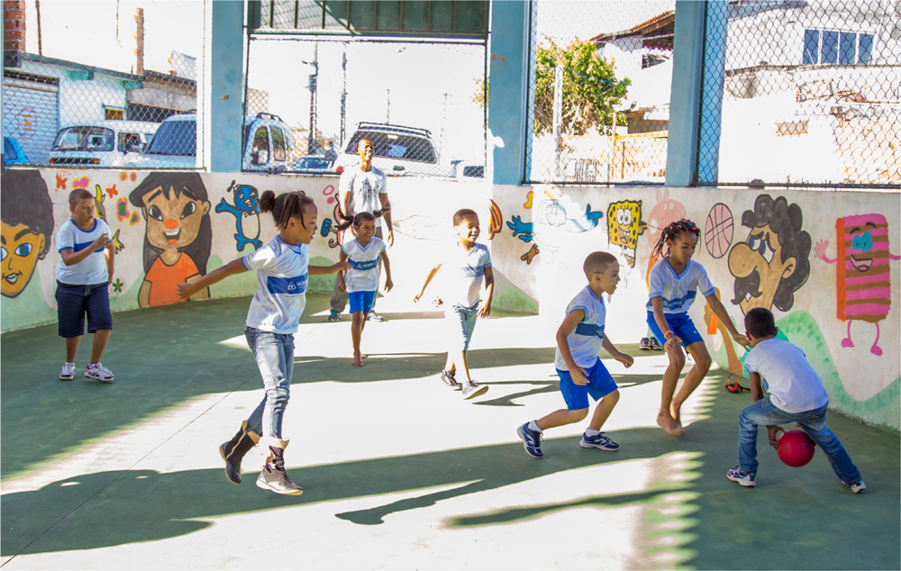 Crianças correm em direção a um menino que está segurando a bola.