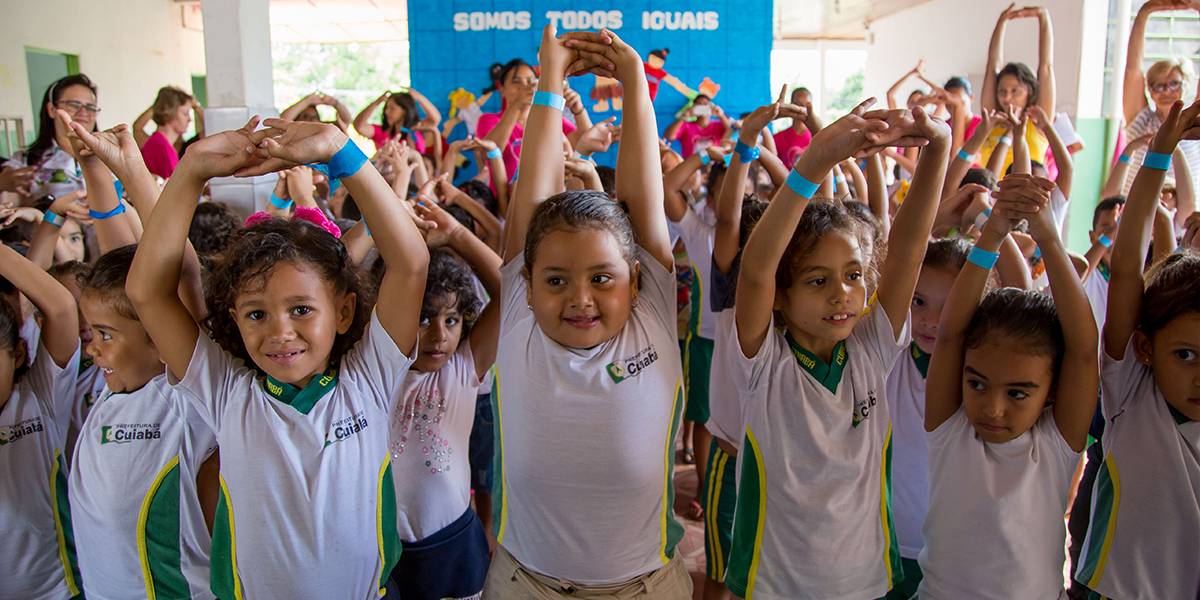 Crianças fazem alongamento esticando os braços para cima e entrelaçando os dedos das mãos sobre a cabeça em um pátio escolar.