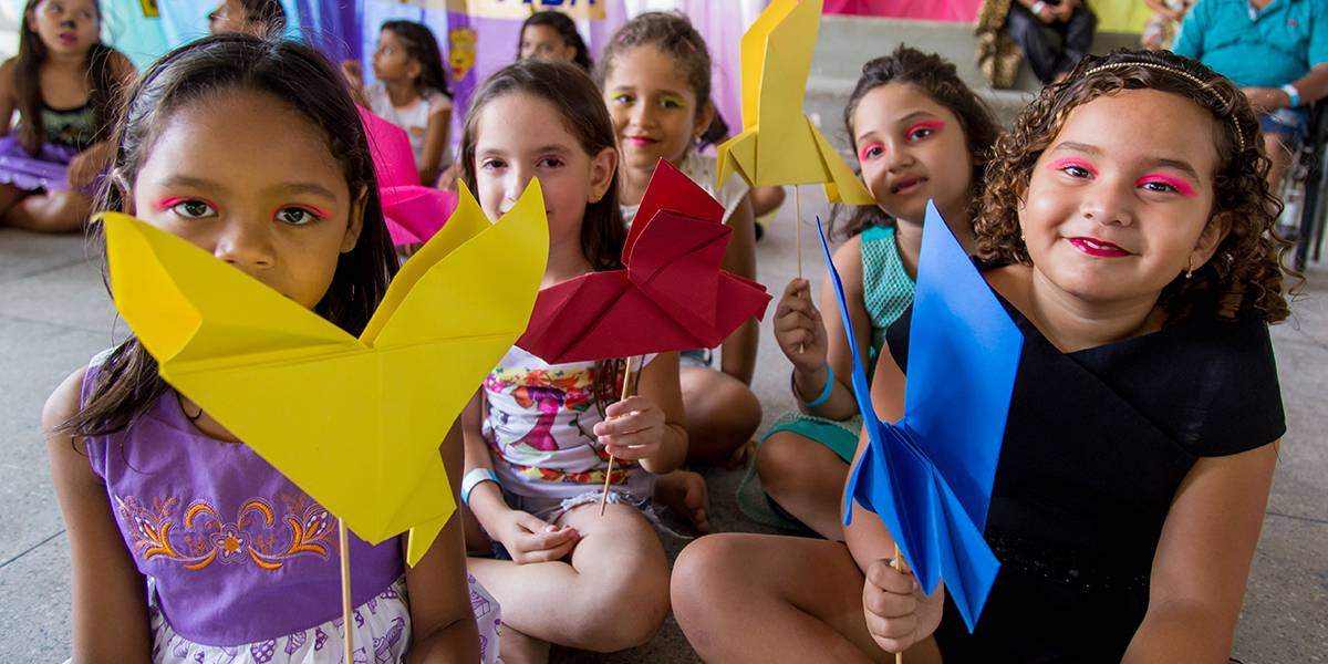 Cinco meninas maquiadas e sentadas no chão posam sorridentes segurando pássaros feitos em origami.