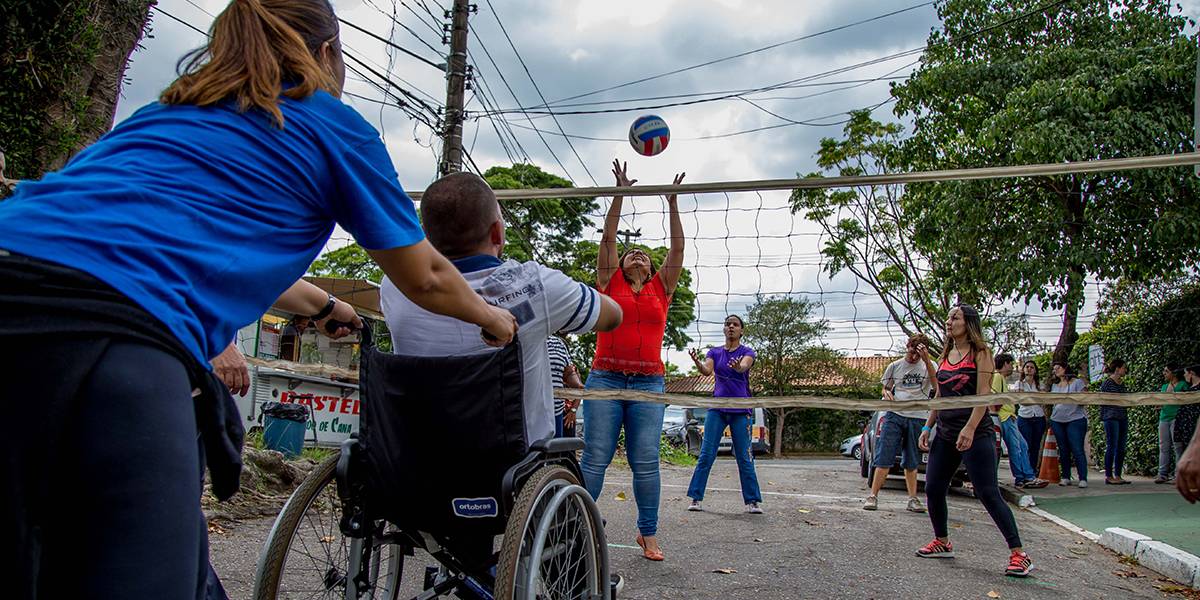 Professora guia cadeira de rodas de homem durante partida de vôlei. Do outro lado da rede, mulher estica os braços para pegar bola que vem em sua direção.