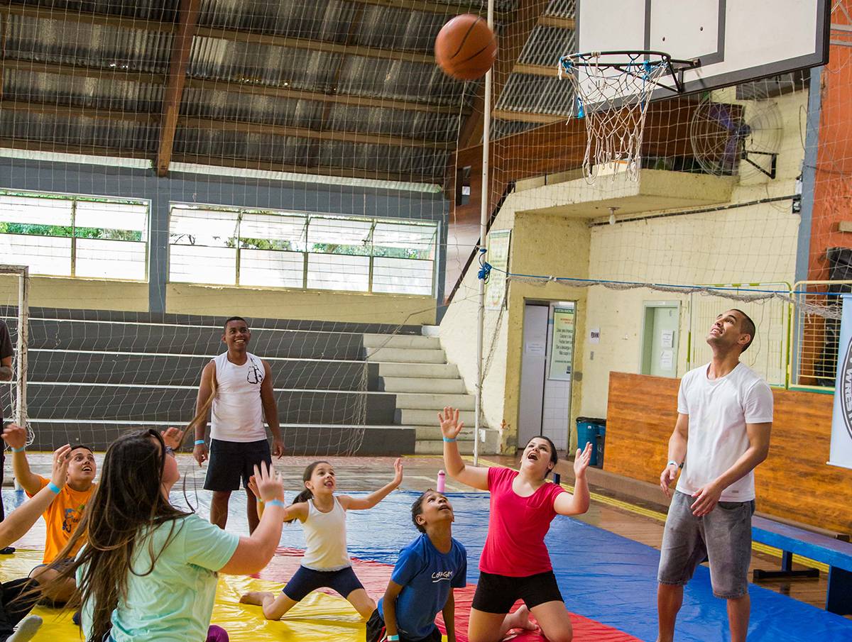 Adolescentes e adultos jogam basquete ajoelhados em chão acolchoado. Com as cabeças para cima, eles acompanham o movimento de uma bola lançada para a cesta.