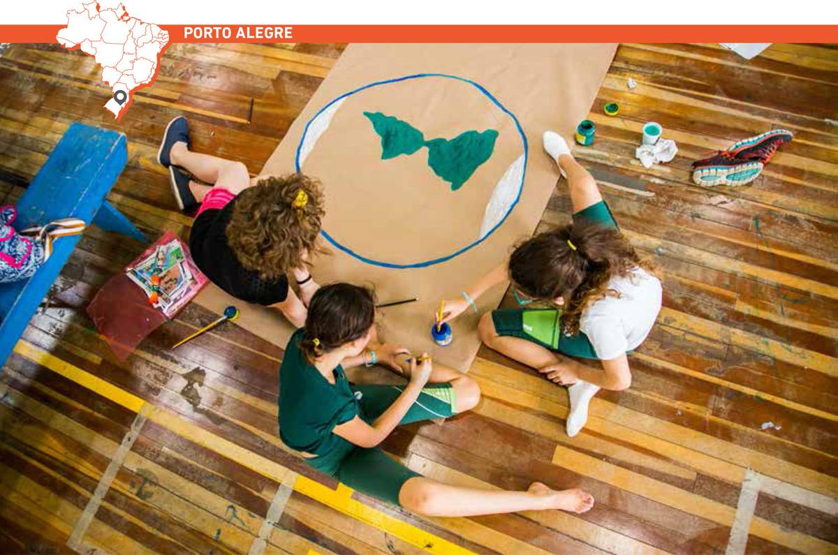 Três garotas sentadas seguram pincéis e potes de tinta para pintar desenho do planeta Terra em grande papel estendido no chão de madeira. Acima da imagem, mapa do Brasil com marcação que destaca cidade de Porto Alegre.