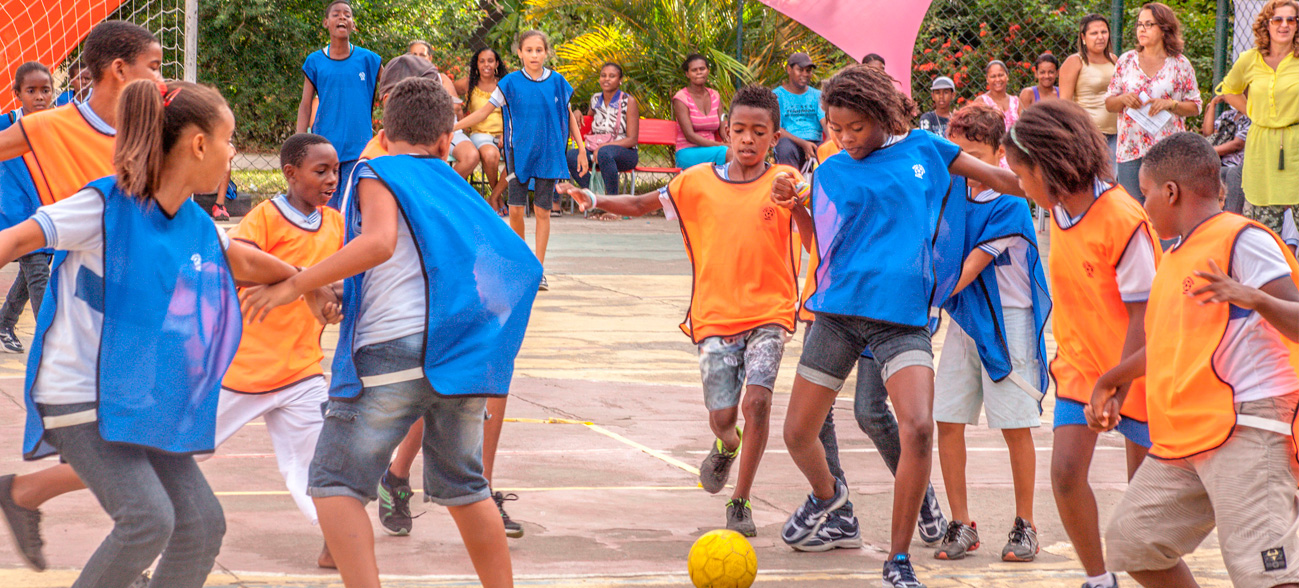 Em uma quadra, meninas e meninos jogam uma atividade com bola. Eles usam coletes de cores diferentes para definir dois times, azul e laranja e correm em pares e de mãos dadas.