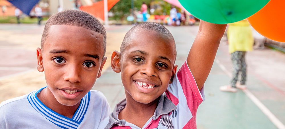 Dois meninos lado a lado numa quadra da escola. Um dele está sorridente e ergue uma das mãos segurando balões coloridos.