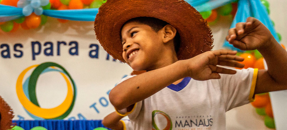 Menino sorri movimentando seus braços para o alto. Ele usa chapéu de palha e uniforme da rede municipal de Manaus.