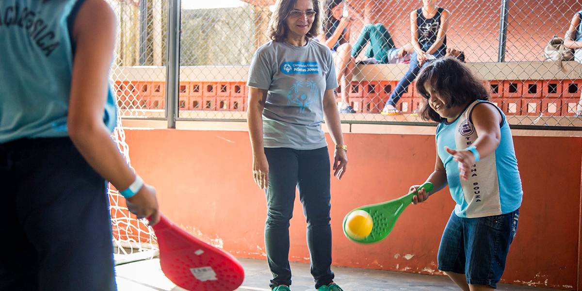 Menina rebate bolinha para o colega com raquete de plástico enquanto professora assiste ao fundo.