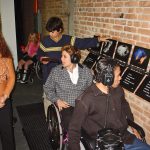 Pessoas com e sem deficiência visitam exposição de arte com recursos de acessibilidade. Visitantes com deficiência física utilizam fone para conferir audiodescrição das obras expostas em parede de tijolo.