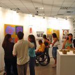 Pessoas com e sem deficiência visitam exposição de arte dentro de uma sala branca. Elas observam atentamente quadros e esculturas, destacadas por focos de luz.
