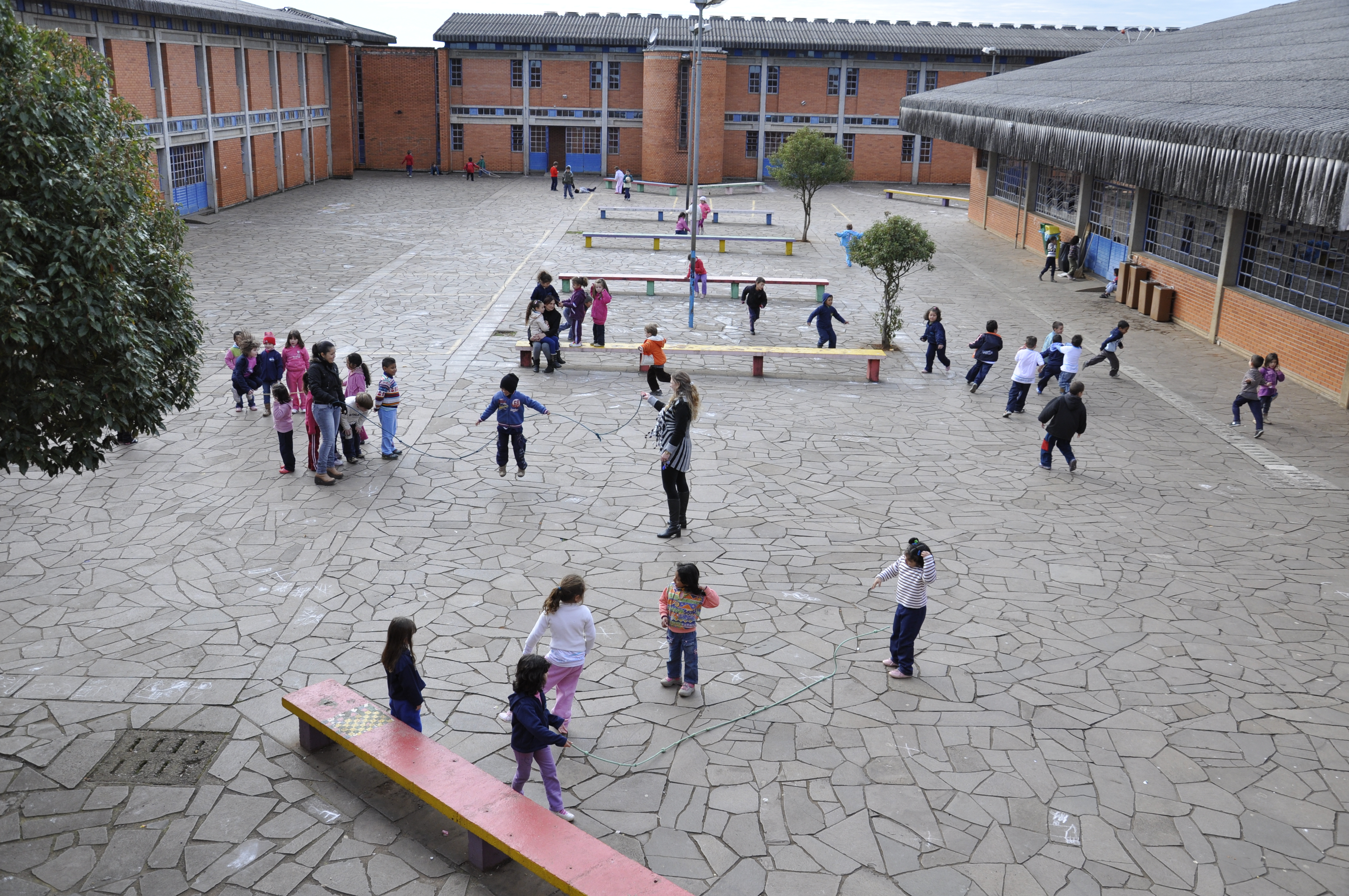 Em pátio amplo escolar, crianças brincam de pular corda e pega-pega divididas em grupos. O dia está nublado e no espaço há bancos compridos e coloridos.