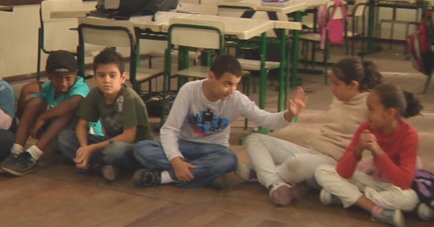 Crianças sentadas lado a lado de pernas cruzadas dentro de sala de aula. Um dos garotos acena para a colega ao seu lado. Ao fundo, mesas e cadeiras escolares.