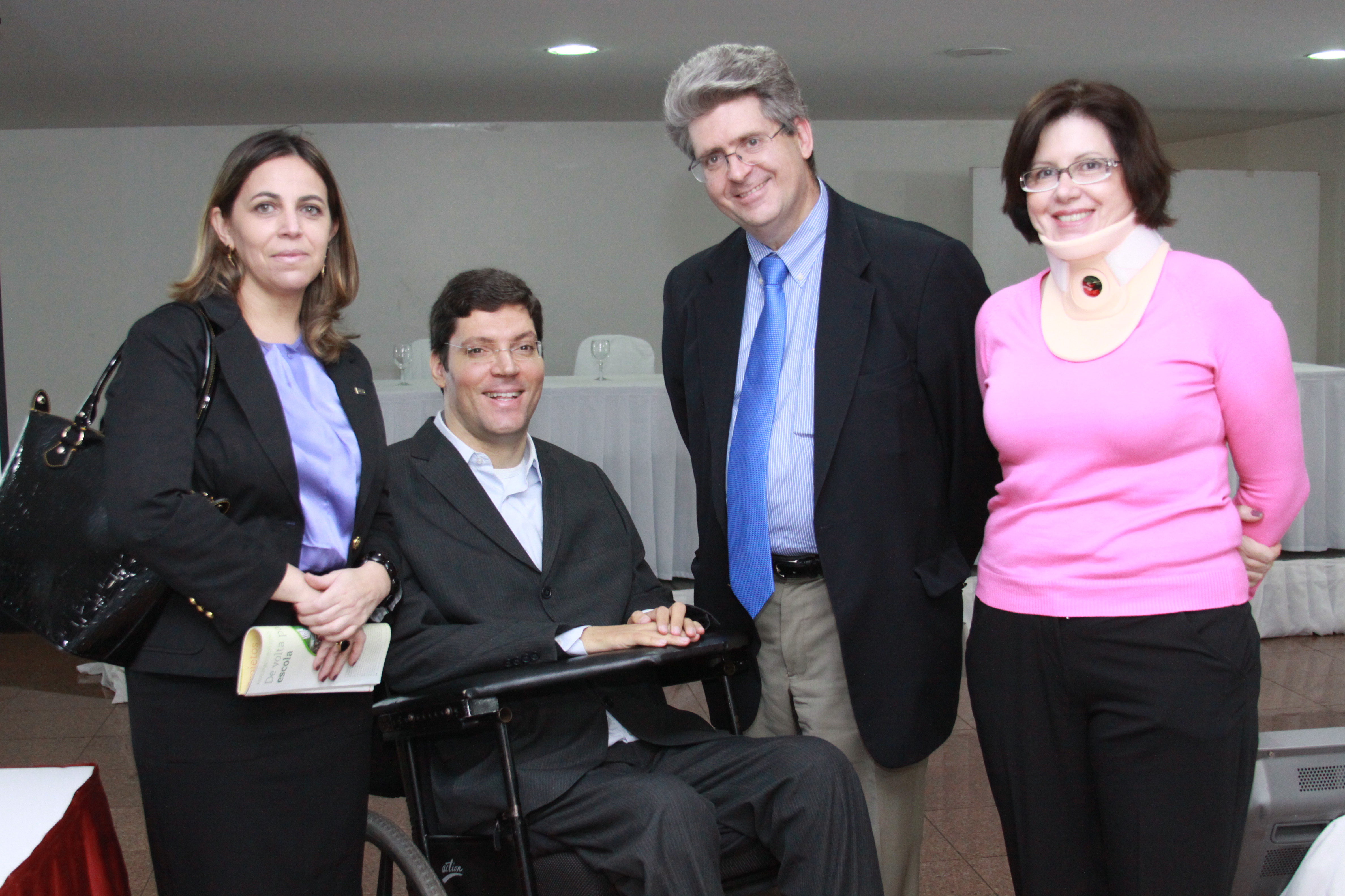 Em destaque, duas mulheres e dois homens em trajes sociais posam lado a lado sorridentes para a foto. Rodrigo está em sua cadeira de rodas e as demais pessoas estão em pé.