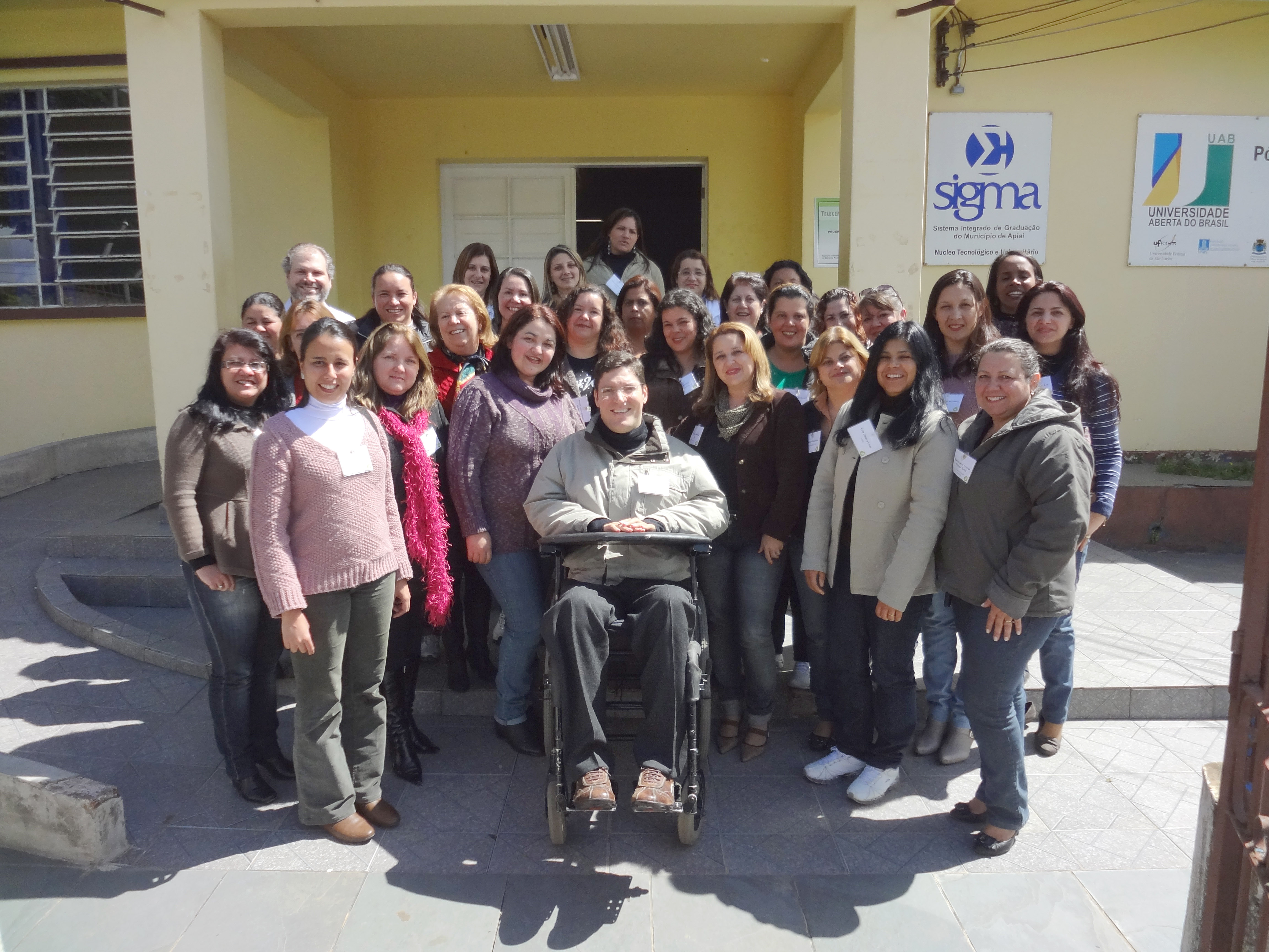 Grupo de educadoras posam sorridentes para foto em frente à universidade junto ao Rodrigo. O dia está ensolarado.