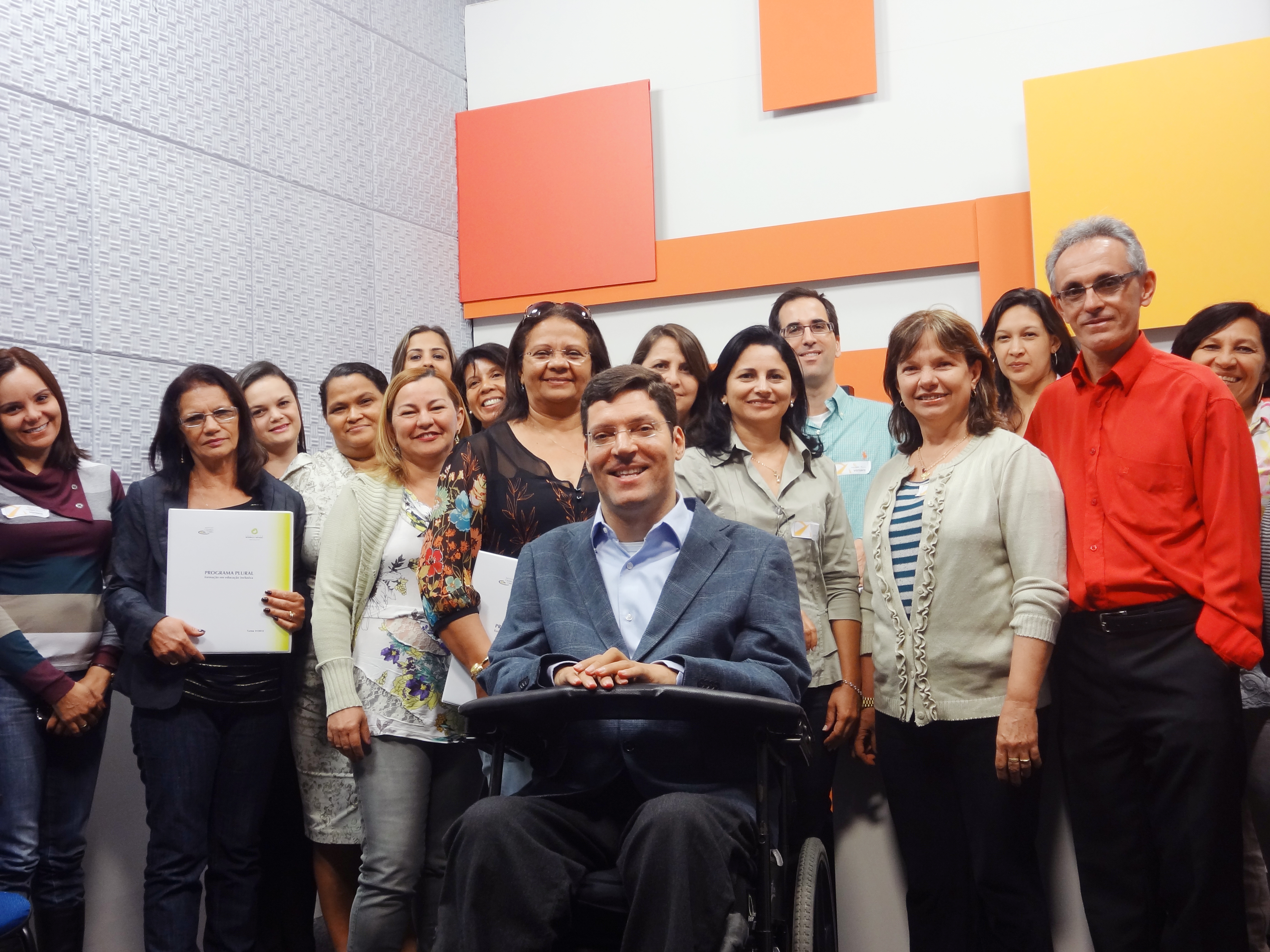 Mulheres e homens posam sorridentes e enfileirados para foto dentro de sala com paredes coloridas. À frente deles está Rodrigo em sua cadeira de rodas.