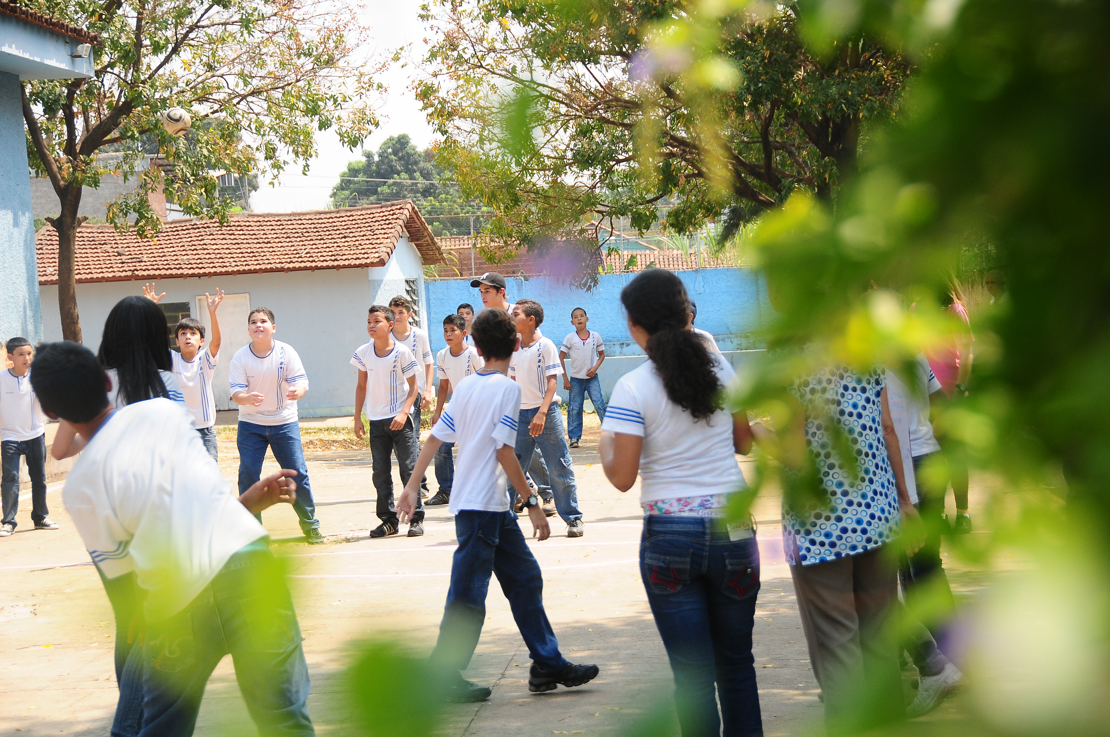 Garotos e garotas uniformizados brincam com bola em pátio do colégio em dia ensolarado.