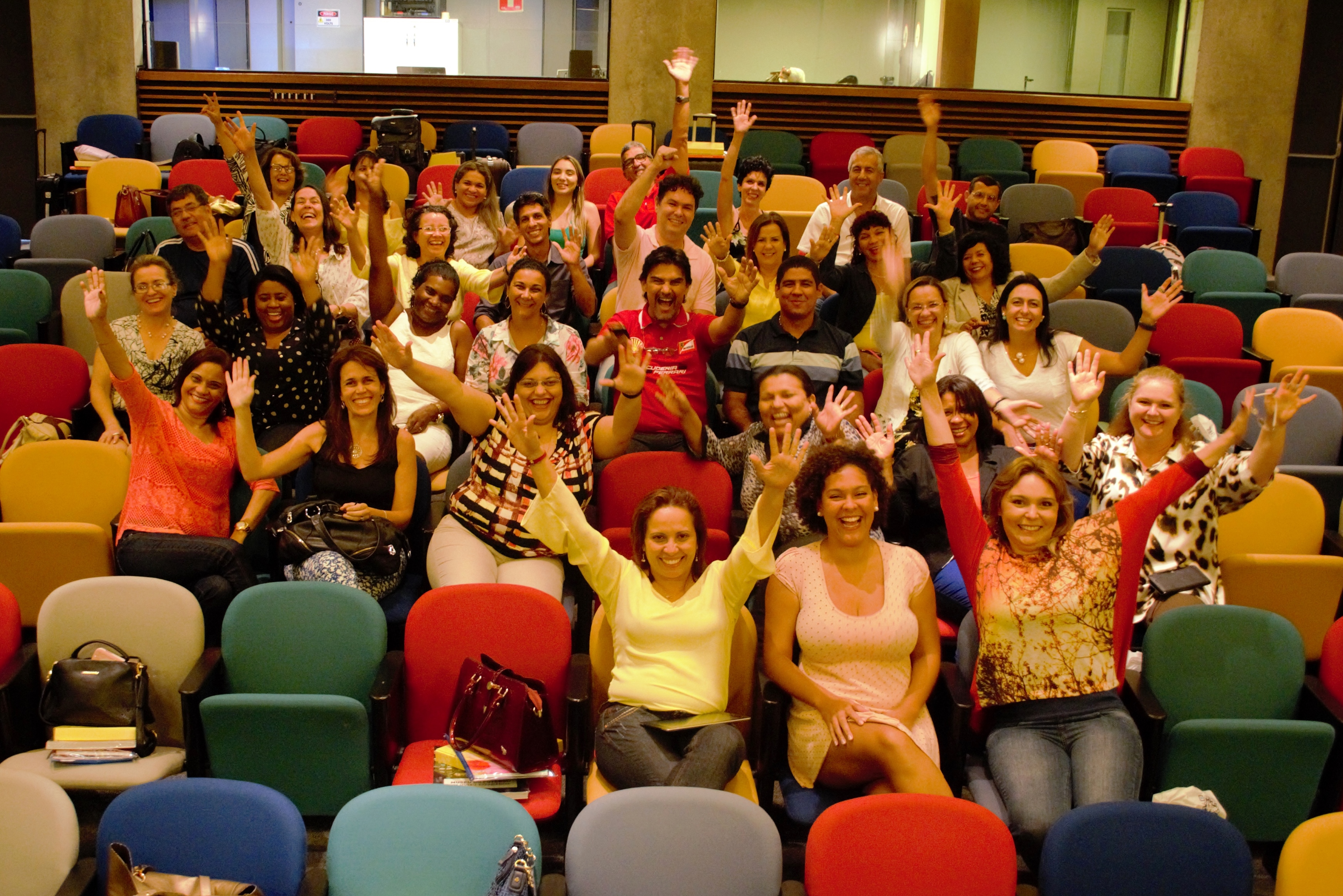 Homens e mulheres com os braços erguidos posam animados para foto em auditório com cadeiras coloridas.