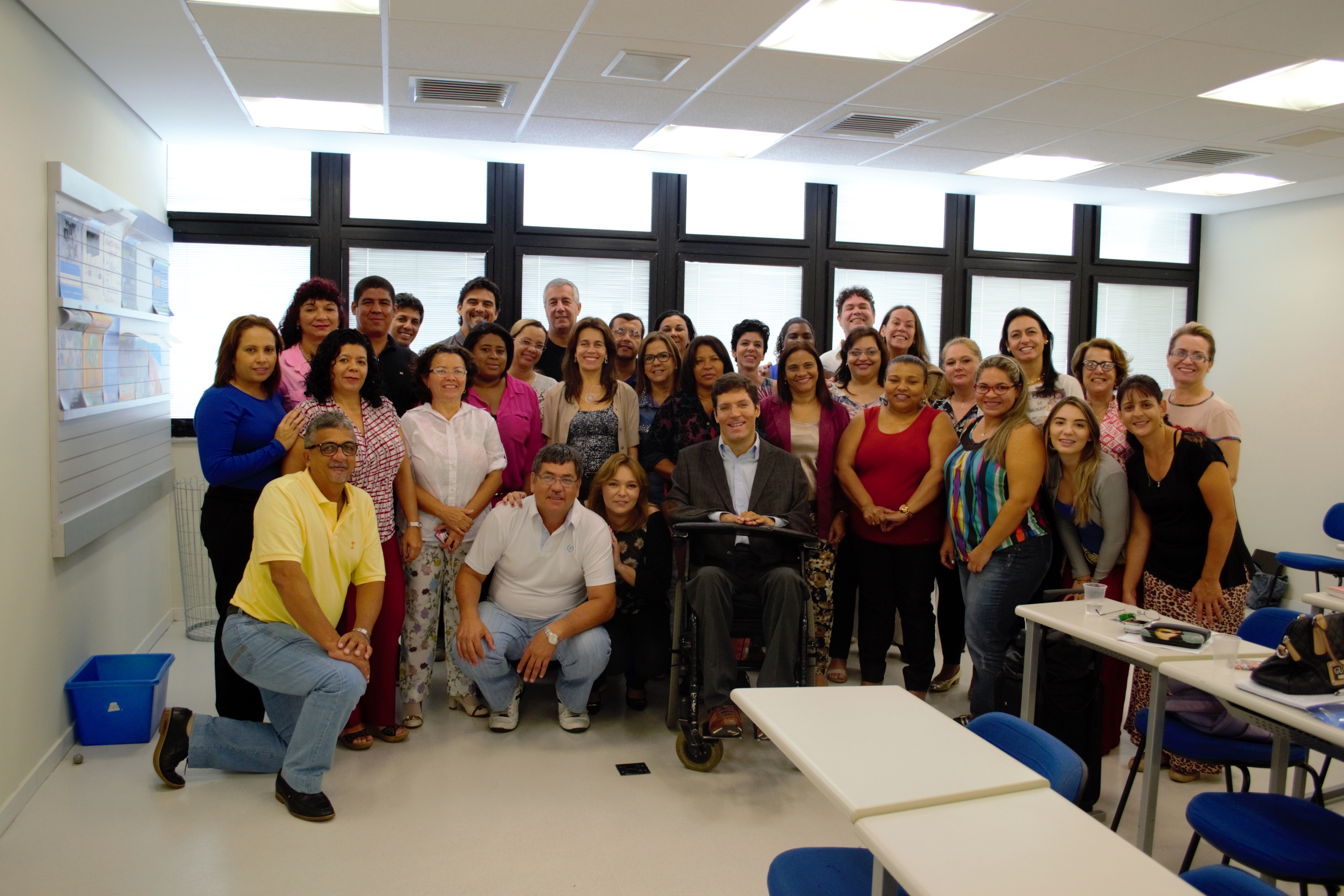 Homens e mulheres posam para a foto agrupados próximos ao Rodrigo, que está no centro do grupo, em cadeira de rodas. Ao fundo, as janelas da sala de aula.