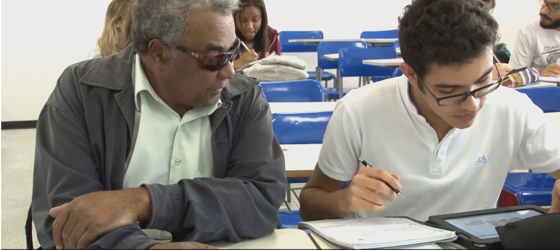 Homem idoso com deficiência visual presta atenção em jovem sentado ao lado direito, que manuseia caderno e tablet. Eles estão em uma sala de aula, sentados lado a lado em mesas escolares.