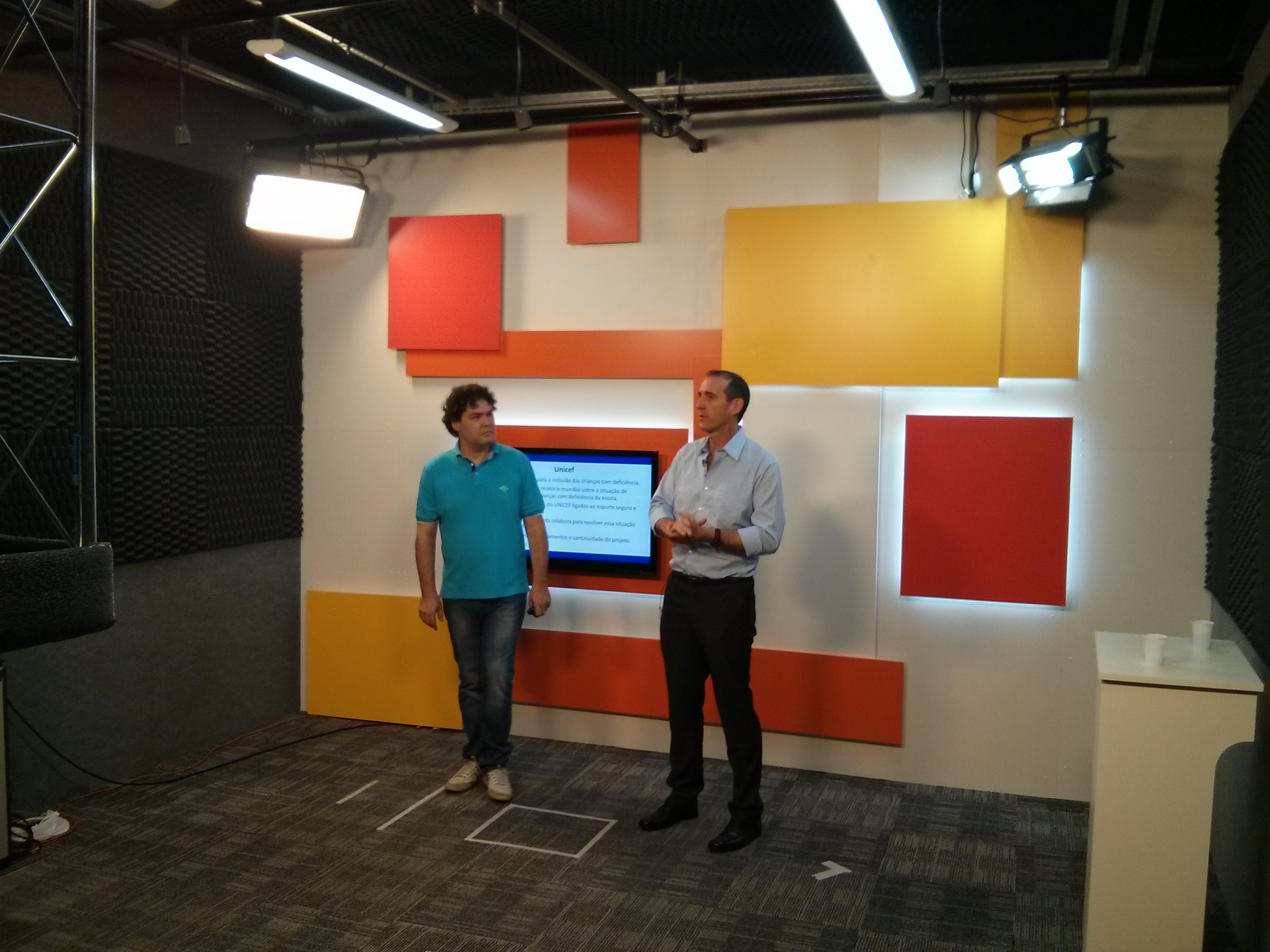 Em estúdio, dois homens estão em pé, a frente de uma parede com quadrados coloridos. Na parede há uma televisão e dois refletores posicionados nos cantos superiores.
