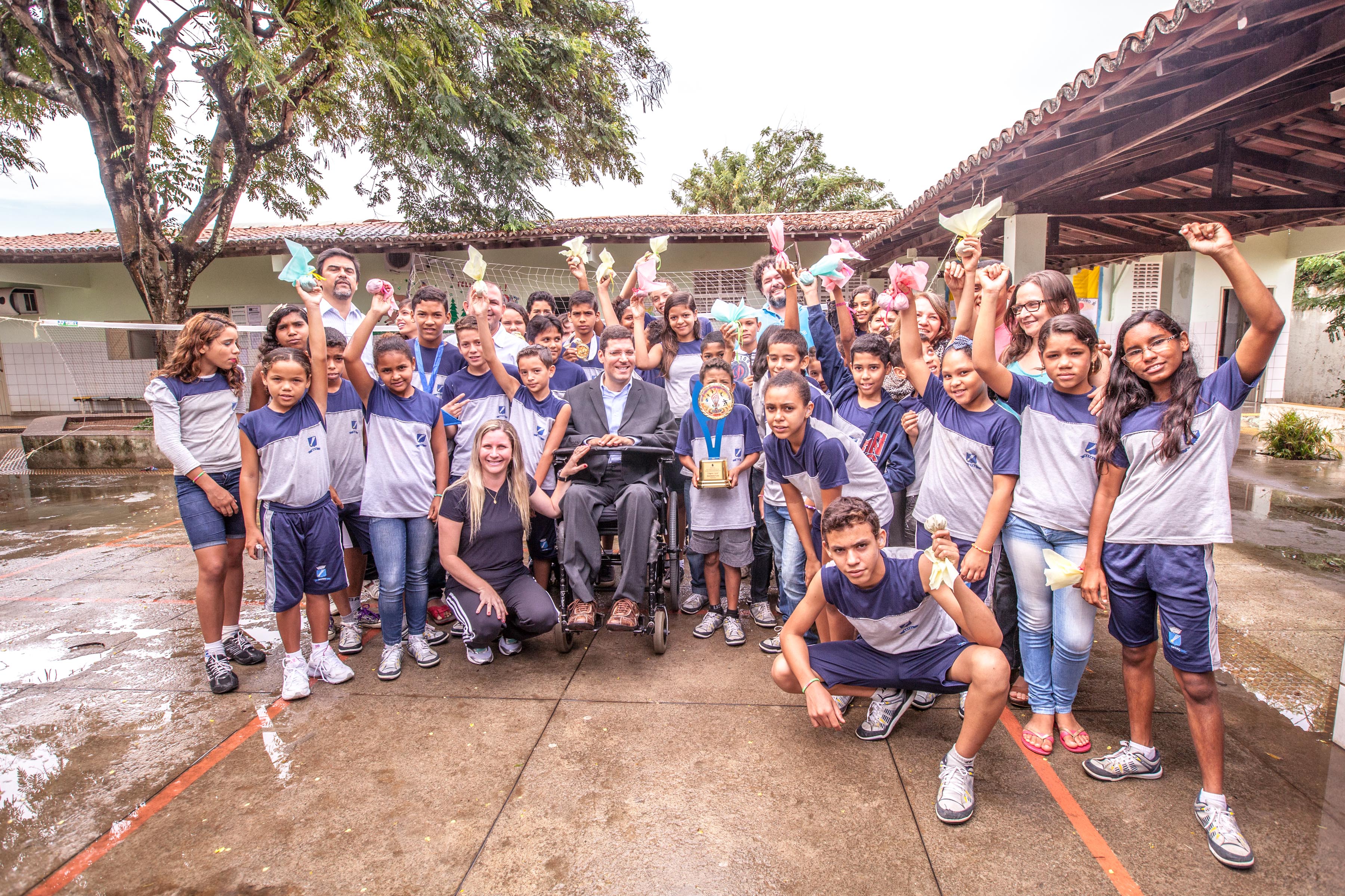 Crianças uniformizadas e educadores posam para foto junto a Rodrigo em pátio de escola. Todos estão sorridentes e alguns estão com os braços erguidos segurando petecas.