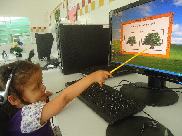 Menina sentada a frente de computador aponta com vareta para a tela onde há figuras de duas árvores, sendo uma maior que outra. Ela está concentrada na atividade e usa fones de ouvido.