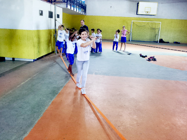 Em quadra poliesportiva, três crianças caminham sobre fita laranja esticada enquanto seguram-se em corda na altura de seus ombros. Ao fundo, outras crianças aguardam em fila junto ao professor.