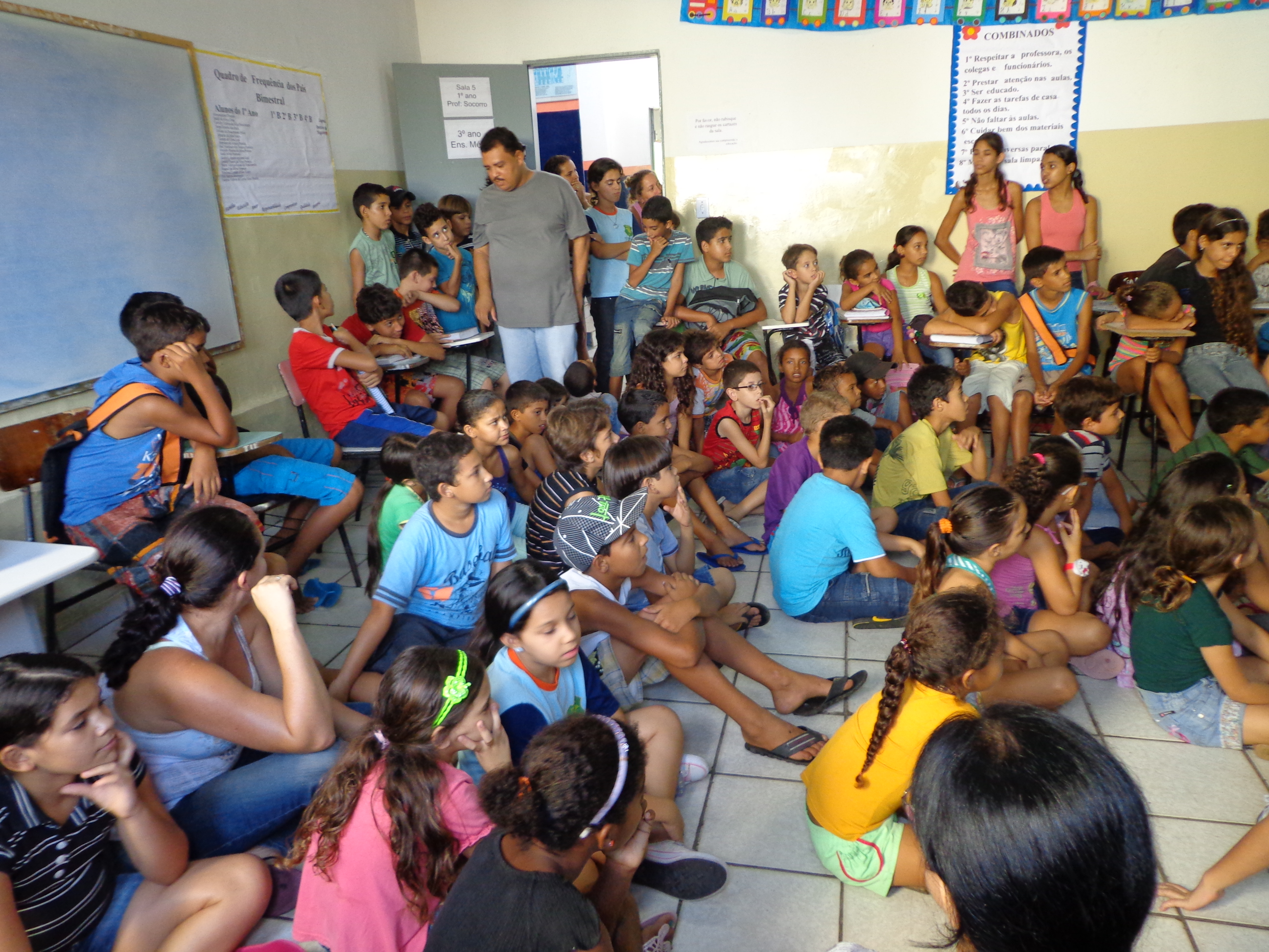 Crianças e adolescentes sentados no chão e cadeiras encostadas na parede da sala de aula. Eles estão voltados para a lousa.