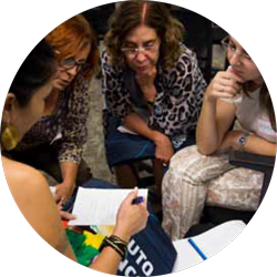 Mulheres sentadas em círculo olham atentamente para folha de caderno posicionada no centro do grupo