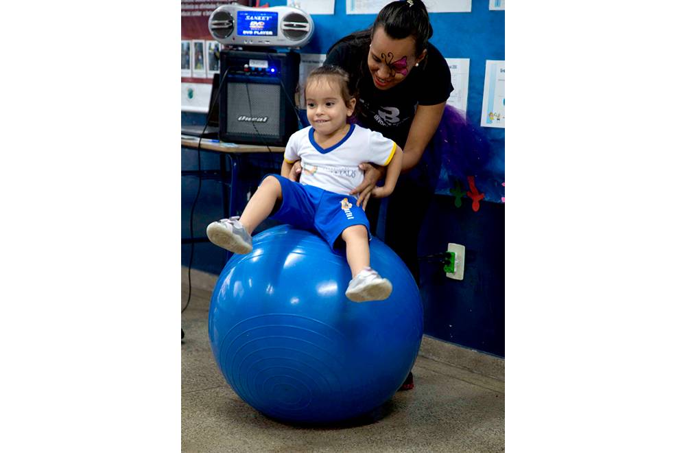 Menina sentada em uma grande bola azul, enquanto professora a segura por trás.