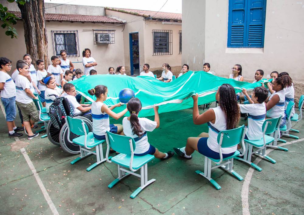 Em pátio escolar, estudantes seguram extremidades do tecido que simula um campo de futebol