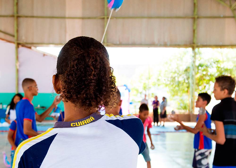 Menina com o uniforme escrito Cuiabá brinca com amigos em quadra escolar.
