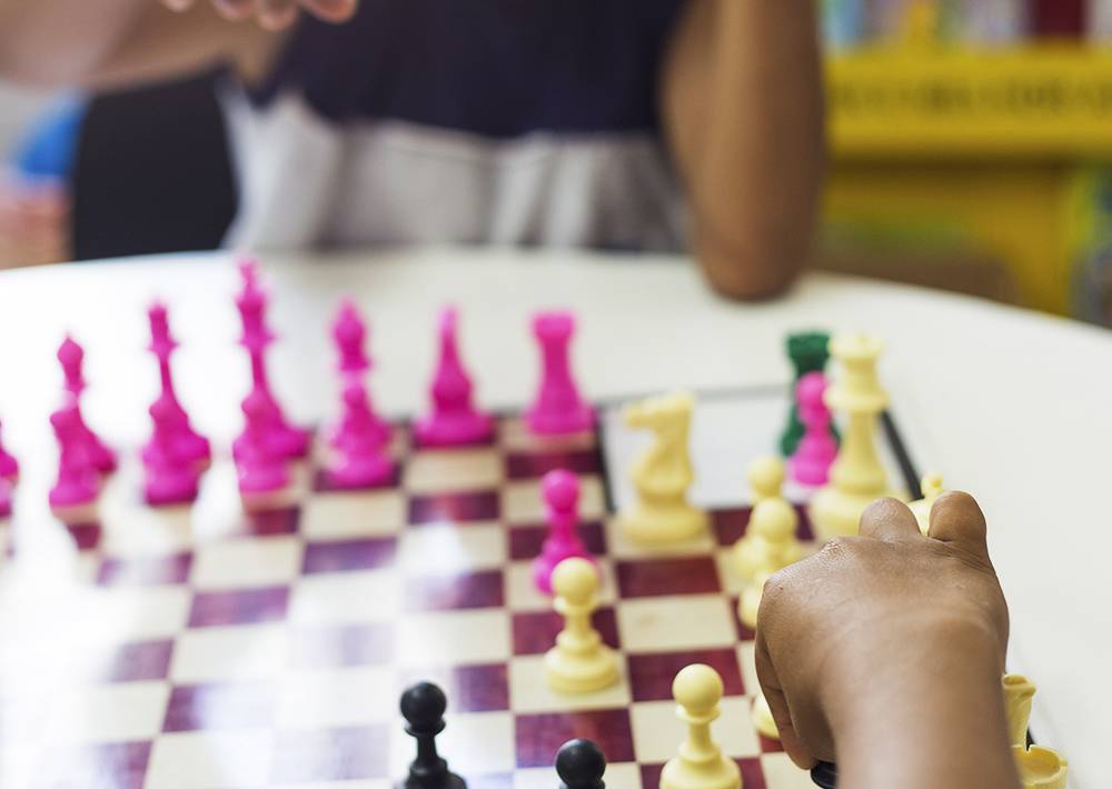 Criança movimenta peça de xadrez e enquanto colega aguarda sua jogada
