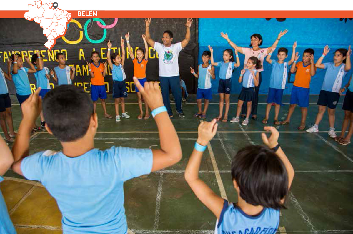 Em roda numa quadra de esportes, professor orienta grupo de estudantes que se alonga esticando os braços para cima. Acima da imagem, mapa do Brasil com marcação que destaca cidade de Belém.