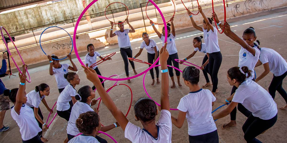 Em círculos, estudantes levantam bambolês sobre suas cabeças durante coreografia.