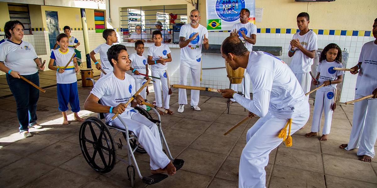Homem joga capoeira com jovem em cadeira de rodas. Ao redor, crianças e adultos batem bastões de madeira.