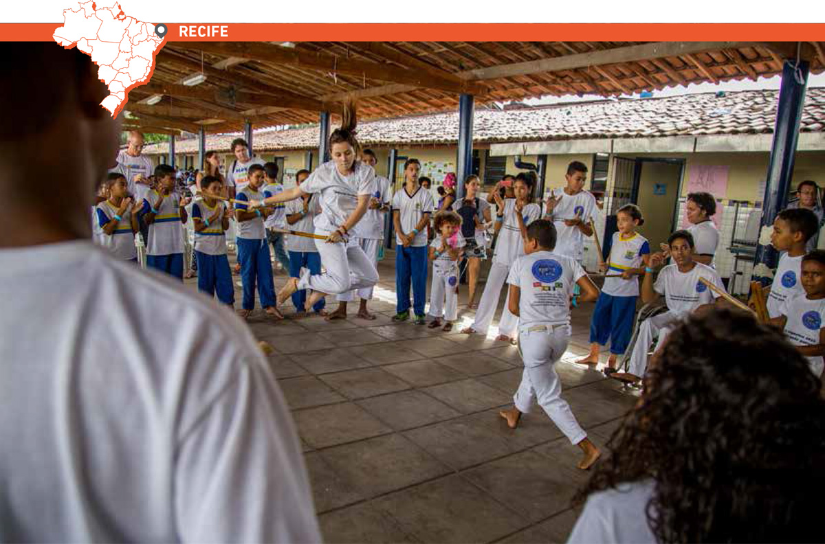 No centro de uma roda de capoeira, garota salta com bastões de madeira na mão enquanto estudante em sua frente ginga. À volta deles, alunos batem palmas e bastões de madeira. Acima da imagem, mapa do Brasil com marcação que destaca cidade de Recife.