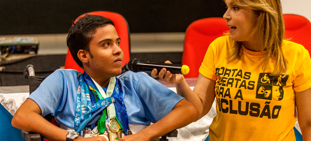 Mulher segura microfone enquanto jovem usuário de cadeira de rodas fala. Ela usa camiseta amarela com os dizeres "Portas abertas para a inclusão". O jovem tem vários cordões com medalhas pendurados em seu pescoço.