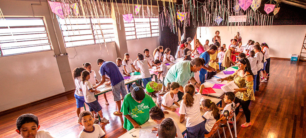 Em uma sala espaçosa há duas grandes mesas, onde crianças orientadas por professores fazem pipas. O teto está enfeitado com rabiolas e pipas coloridas penduradas.