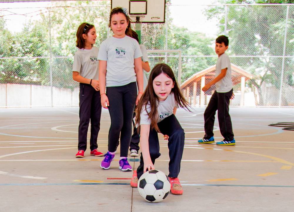 Crianças formam fila em quadra escolar. A primeira da fila está posicionando com a mão uma bola de futebol no chão.