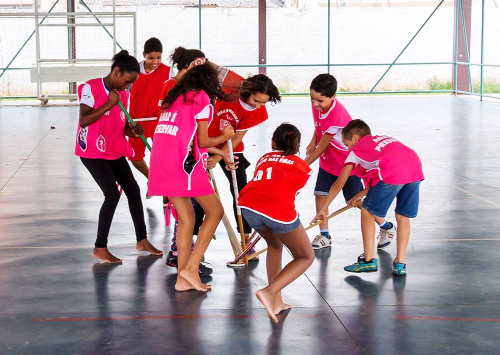 Em uma quadra de esportes, grupo de crianças disputam disco de hóquei no chão com bastões feitos com cabos de vassoura.