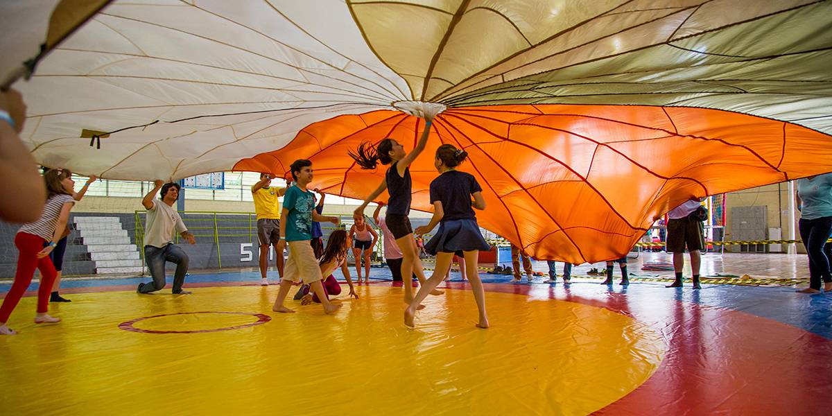 Crianças e jovens sacodem um paraquedas aberto, enquanto outras brincam embaixo do dele. O tecido tem forma circular e quando cai forma uma cabana.
