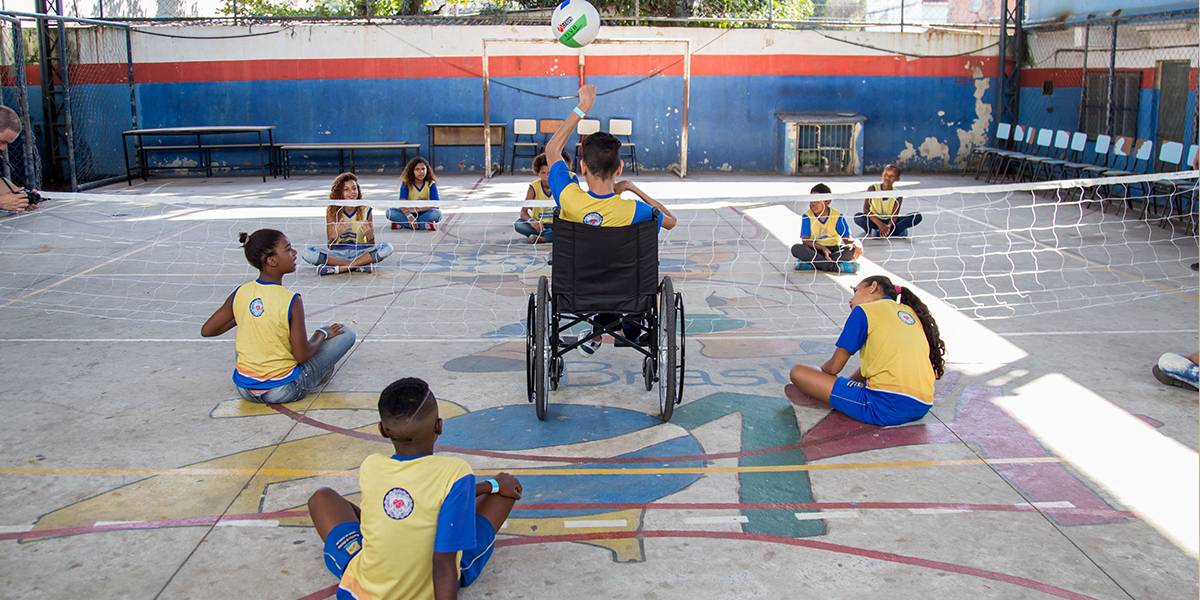 Crianças jogam vôlei sentados na quadra. Ao centro, um menino em cadeira de rodas, se prepara para rebater a bola que chega até ele.
