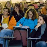 Grupo de mulheres sentadas em auditório em cadeiras universitárias prestam atenção em aula. Algumas delas fazem anotações.