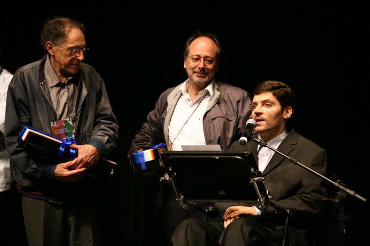 Dois homens em pé seguram presentes e olham com admiração para Rodrigo, enquanto ele fala ao microfone sentado em sua cadeira de rodas.