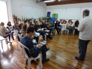 Grupo de pessoas sentadas em cadeiras plásticas que formam um semicírculo. Eles estão fazem anotações atentos ao palestrante que está em pé no centro da sala.