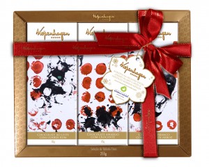 Caixa de embalagem quadrada com borda dourada e a inscrição “Kopenhagen” contendo três tabletes de chocolate em embalagens estampadas com obra de arte abstrata em preto e vermelho. A caixa está envolta por fita vermelha e um grande laço. No laço, cartão com forma de flor com o letreiro “Kopenhagen acredita na inclusão social por meio da arte e apoia o Instituto Rodrigo Mendes”.